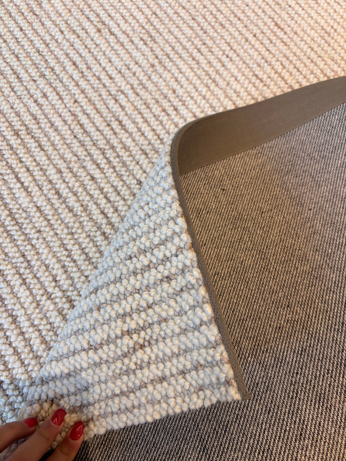 Corner of the Glint rug folded back by hand, showing its ribbed oatmeal loops and the grey backing.