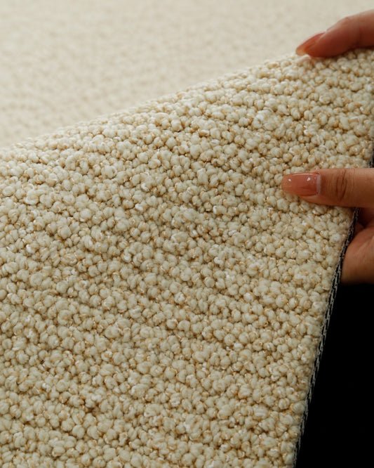 Hands lifting the edge of the Sunlit rug, showing its golden-beige bouclé loops.