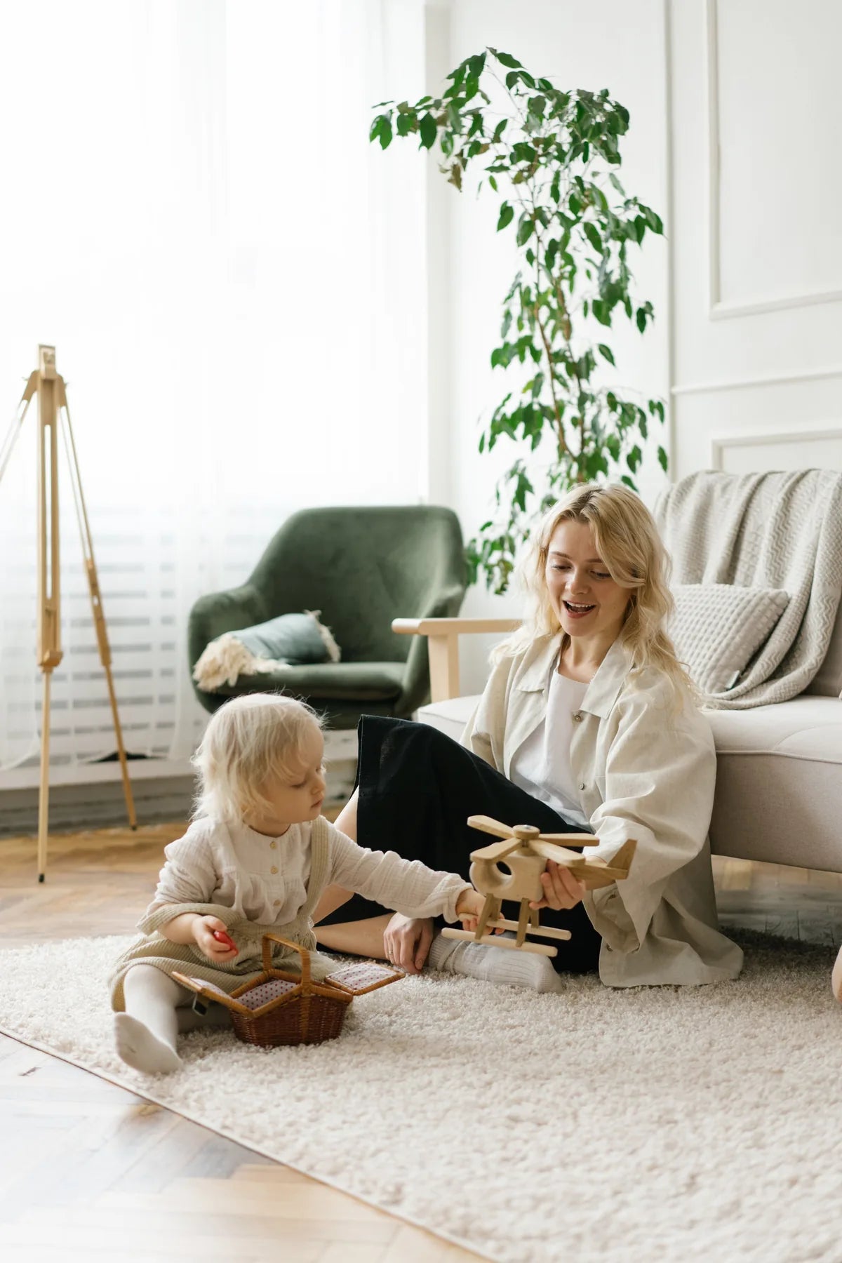 A woman smiles as a toddler plays with a wooden toy on the living-room floor.