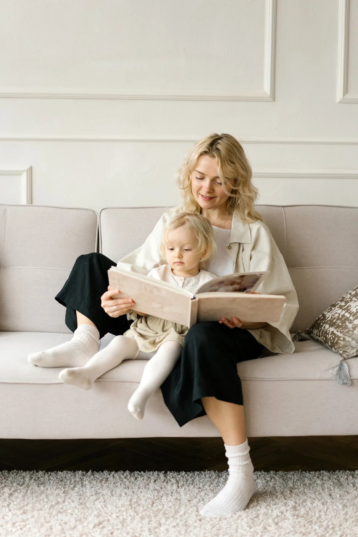 A woman reads a picture book with a small child on her lap, curled up on a pale sofa.