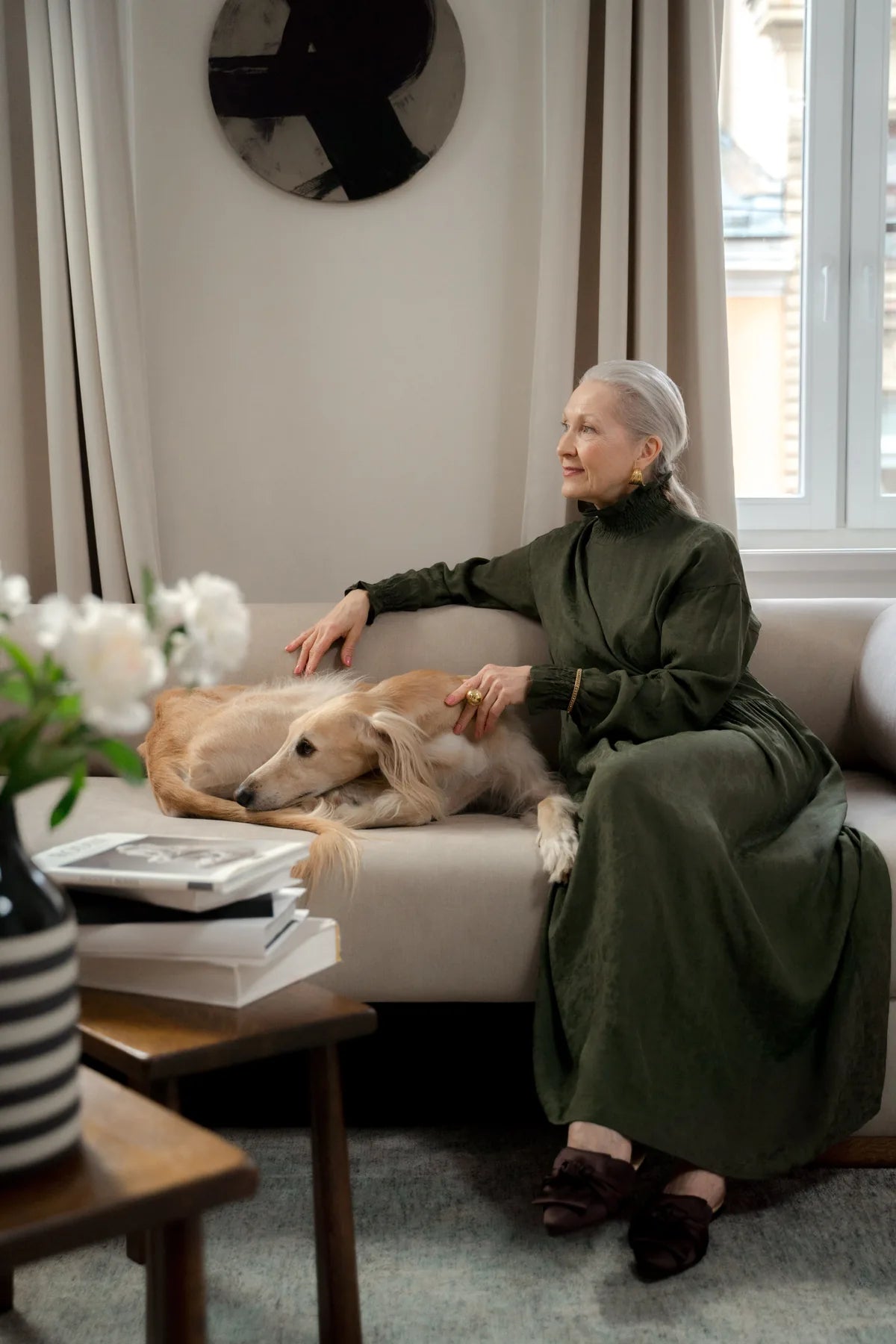 An older woman sits on a sofa beside her resting dog, looking towards the window light.